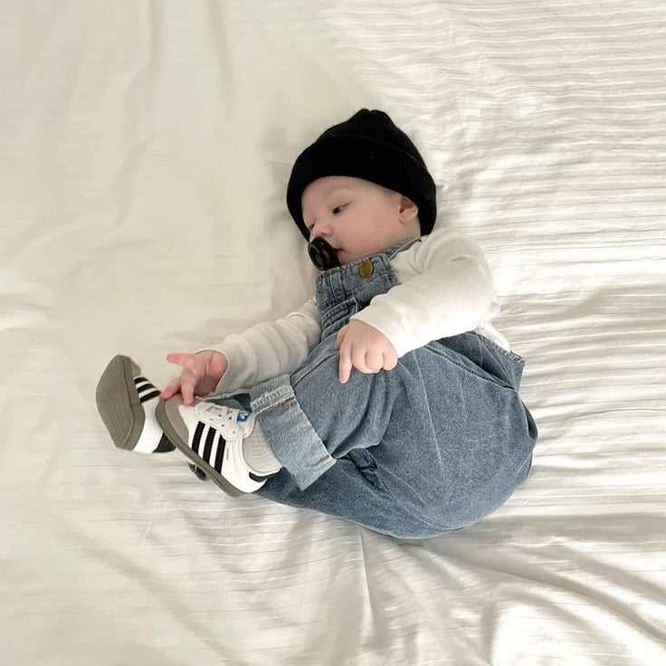 Baby lying on a white bed wearing a black hat, white shirt, blue jeans, and striped socks.